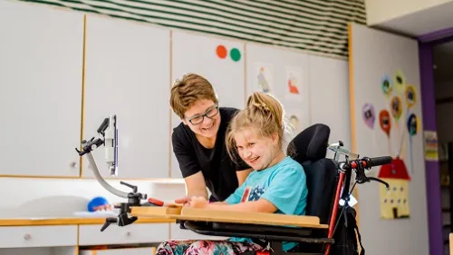Ein Mädchen sitzt im Rollstuhl, daneben steht eine Mitarbeiterin